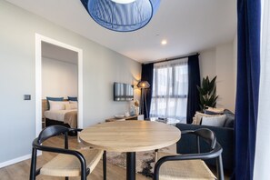 A modern living room with a round wooden table, two chairs, a sofa, a TV mounted on the wall, and a large window with curtains. - Be Casa - Valdebebas (Madrd)