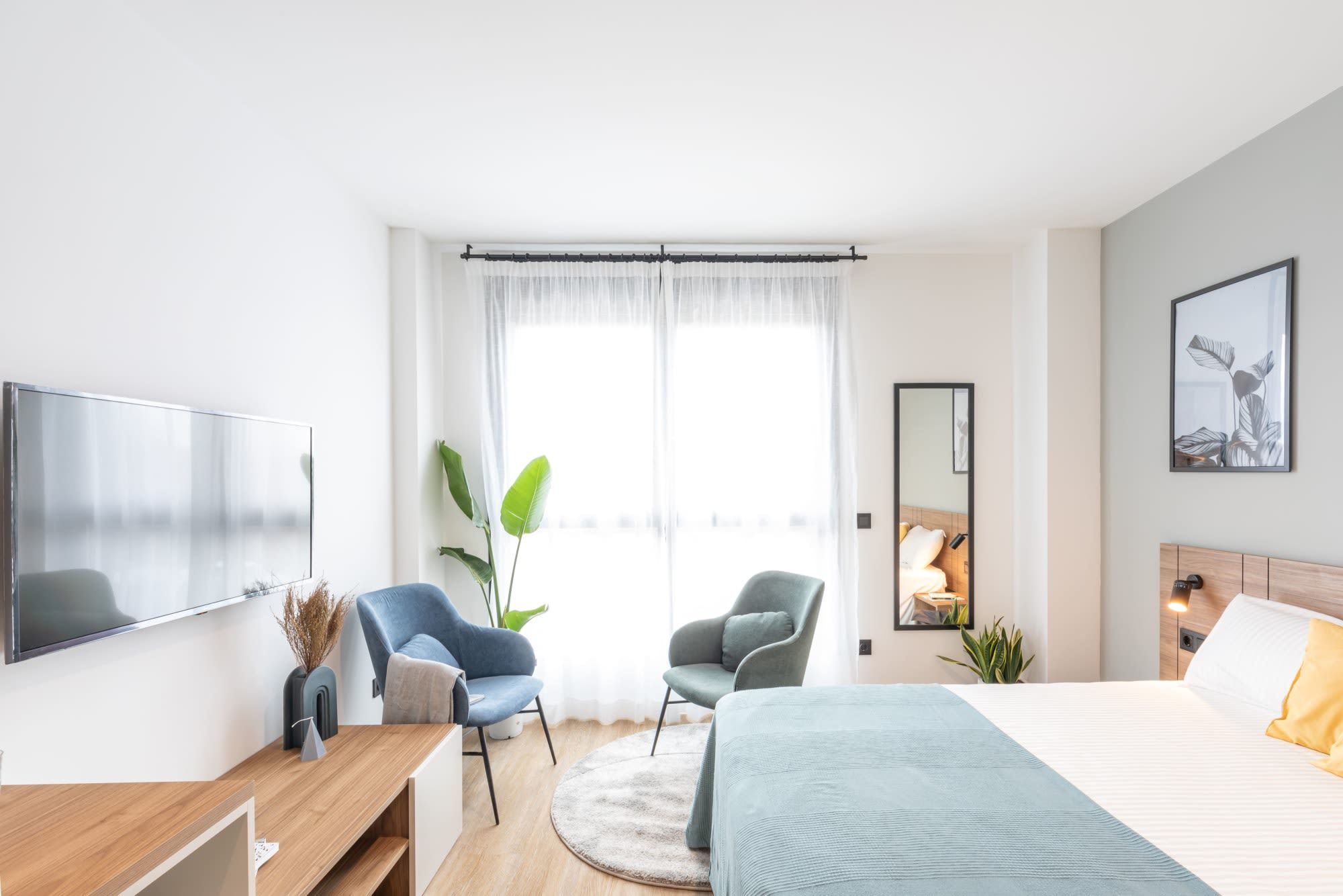 Een moderne hotelkamer met een bed, twee stoelen, een bureau, een flatscreen televisie, een groot raam met dunne gordijnen en een spiegel aan de muur.