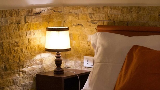 A hotel room with a stone wall, a bedside lamp, a neatly made bed with a green bedspread, and a rolled white towel on the bed. - Vacation House Dolce Angelo 21, with view of Amphitheater, in the heart of the historic center (Sutri)
