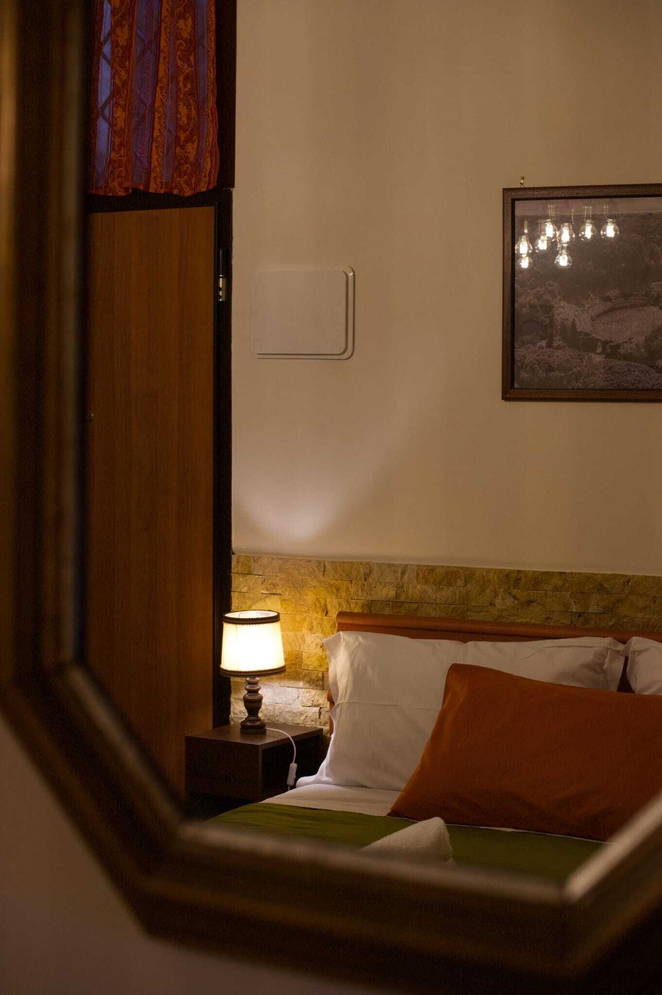 Een hotelkamer met een bed, een nachtkastje met een lamp, een lijst met een afbeelding aan de muur en een raam met gordijnen.
