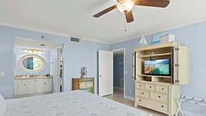 Une chambre à coucher comprenant un lit, un téléviseur, un ventilateur de plafond, une coiffeuse avec un miroir et une porte donnant sur une autre pièce.