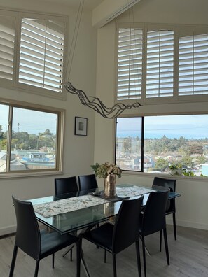 A dining area with a glass table, black chairs, a decorative metal piece, and a view of a residential area through large windows. - Beachfront Bliss Starts Here—Steps to the Sand-3-bedroom house in charming Aptos (Aptos)