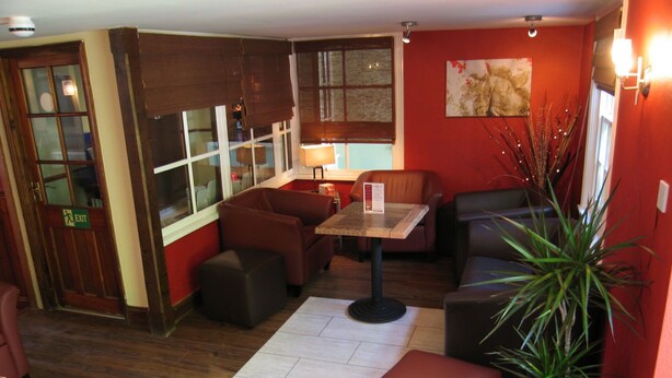 A room with a wooden floor, a table with a lamp, leather sofas, a potted plant, and a red wall. - The George and Dragon Hotel (Sudbury)
