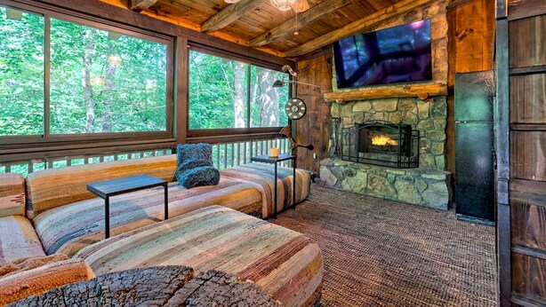 A log cabin interior with a stone fireplace, a large TV mounted above it, and a cozy seating area with a view of the forest.