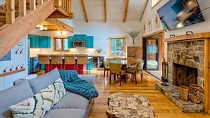 A spacious living area with a high ceiling, wooden beams, a fireplace, a dining area, and a kitchen with teal cabinets.