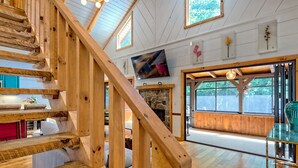 A wooden staircase leading to a living area with a stone fireplace, large windows, and a mounted television.