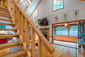 A wooden staircase leading to a living area with a stone fireplace, large windows, and a mounted television.