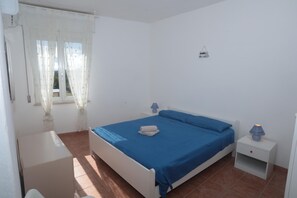 A bedroom with a bed, a nightstand, a small table, and a window with curtains. - Residence Cala di Seta three-room apartment Luca with sea view for 5 people (Calasetta)
