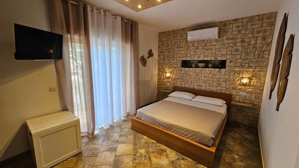 A bedroom with a bed, curtains, a flat-screen TV, a stone-patterned wall, and a mounted air conditioner.