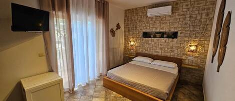 A bedroom with a bed, curtains, a flat-screen TV, a stone-patterned wall, and a mounted air conditioner.
