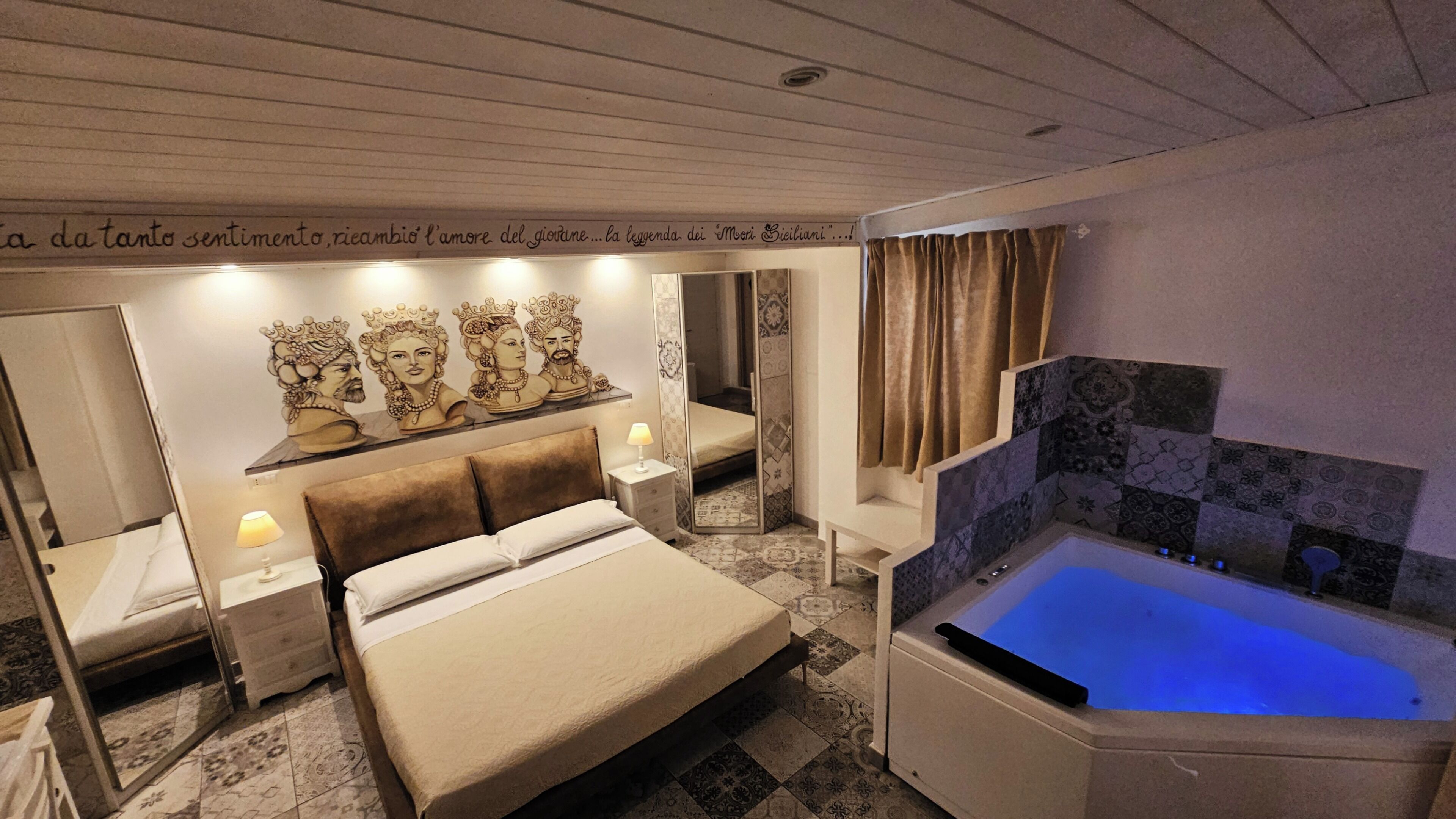 Habitación de hotel con cama, mesita de noche, espejo, un cuadro en la pared y baño con bañera, además de piso estampado.