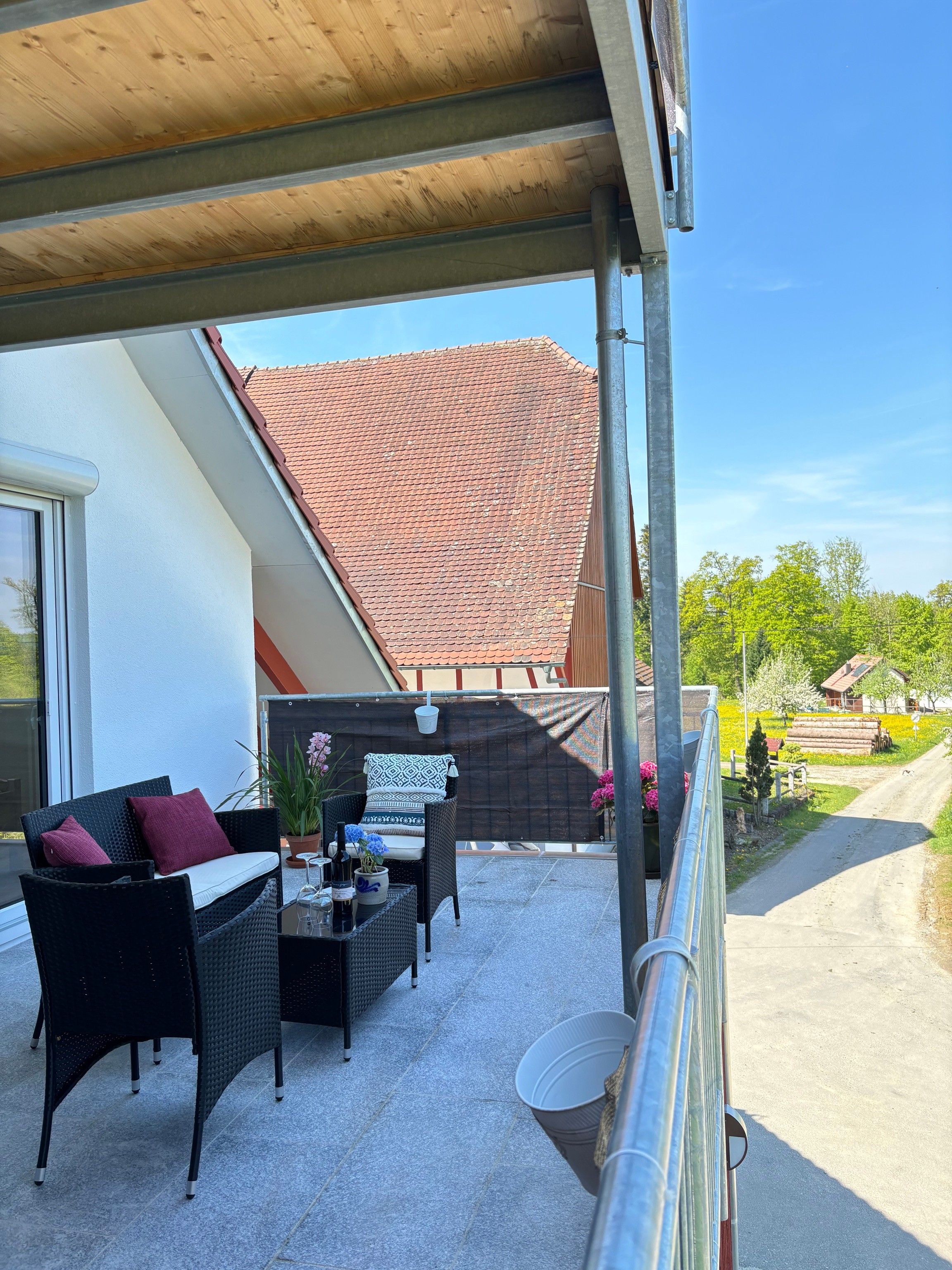 A covered outdoor seating area with wicker furniture, a table with drinks, and a view of a red-tiled roof building.