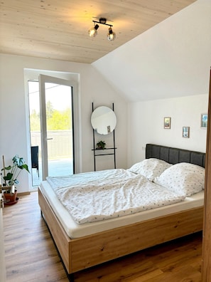 Ein Schlafzimmer mit einem Bett, einer Zimmerpflanze, einem Spiegel und Blick durch eine Tür.
