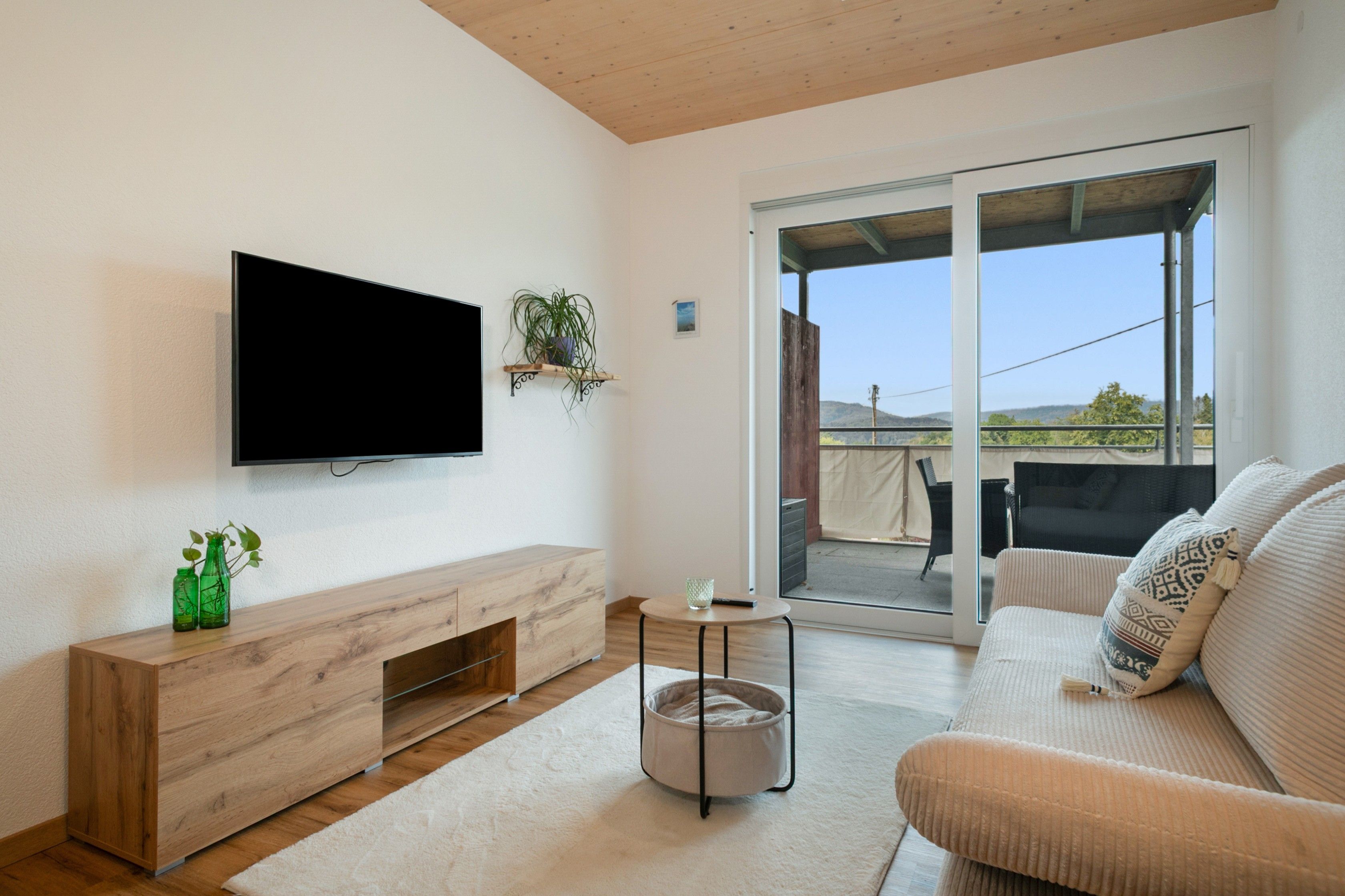 Ein modernes Wohnzimmer mit einem an der Wand montierten Flachbildfernseher, einer Holz-Unterhaltungsecke, einem runden Couchtisch, einer Couch und einer Schiebetür zu einem Außenbereich.