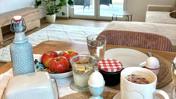 A dining area with a table set for breakfast, including apples, a glass of water, and a cup of coffee. - Panoramic View Holiday Apartment (Deggenhausertal)