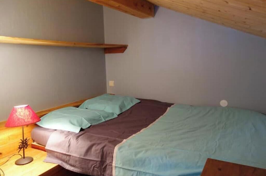 A single bed with a brown and teal bedspread, a red lamp on a wooden nightstand, and a wooden beam above the bed.