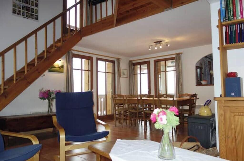 Une pièce avec un escalier en bois, une table à manger avec des chaises, une bibliothèque et un vase rempli de fleurs.