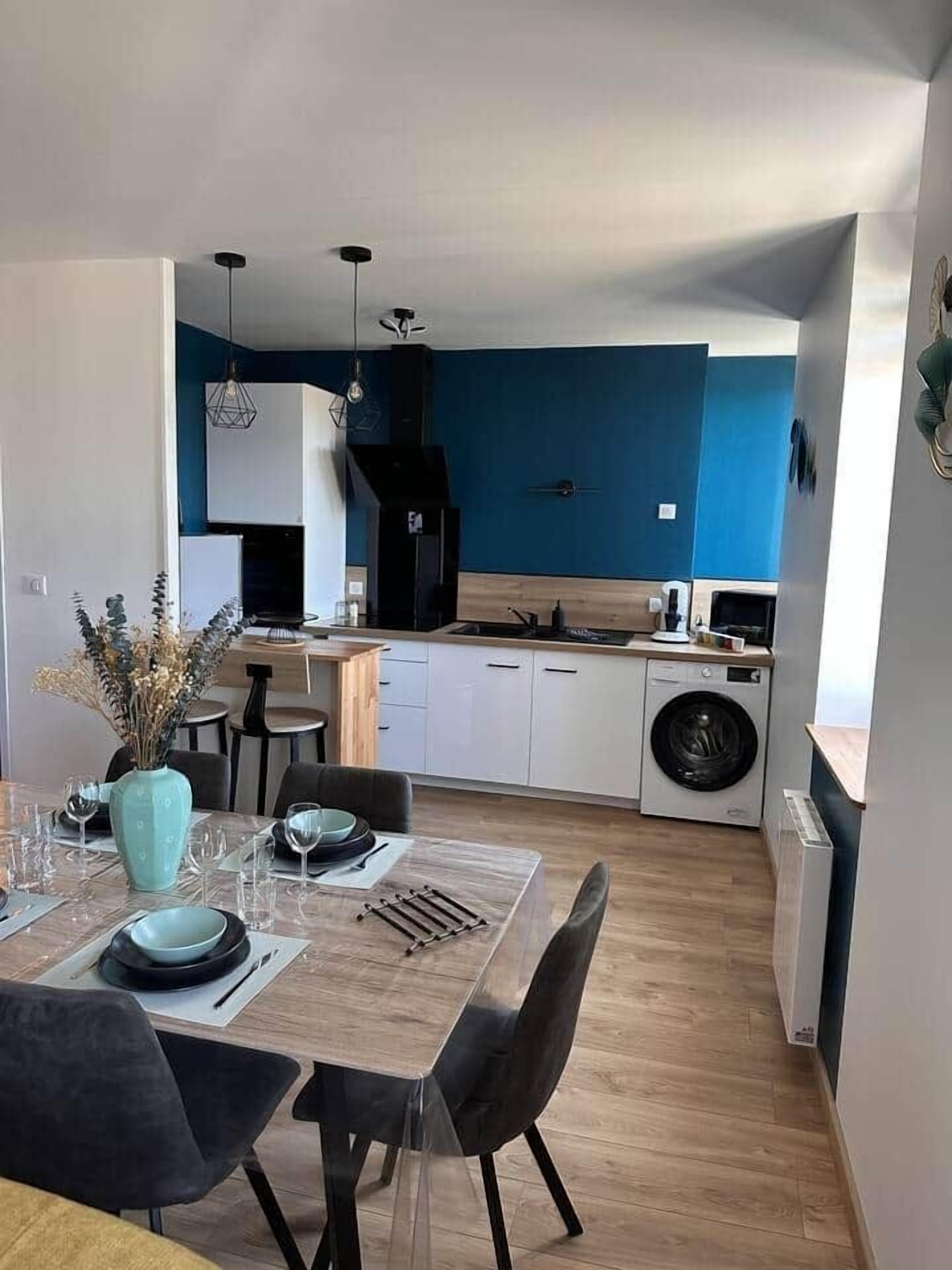 Une cuisine moderne équipée d’un lave-linge, un coin repas avec une table dressée pour deux personnes, et un mur d’accent bleu.