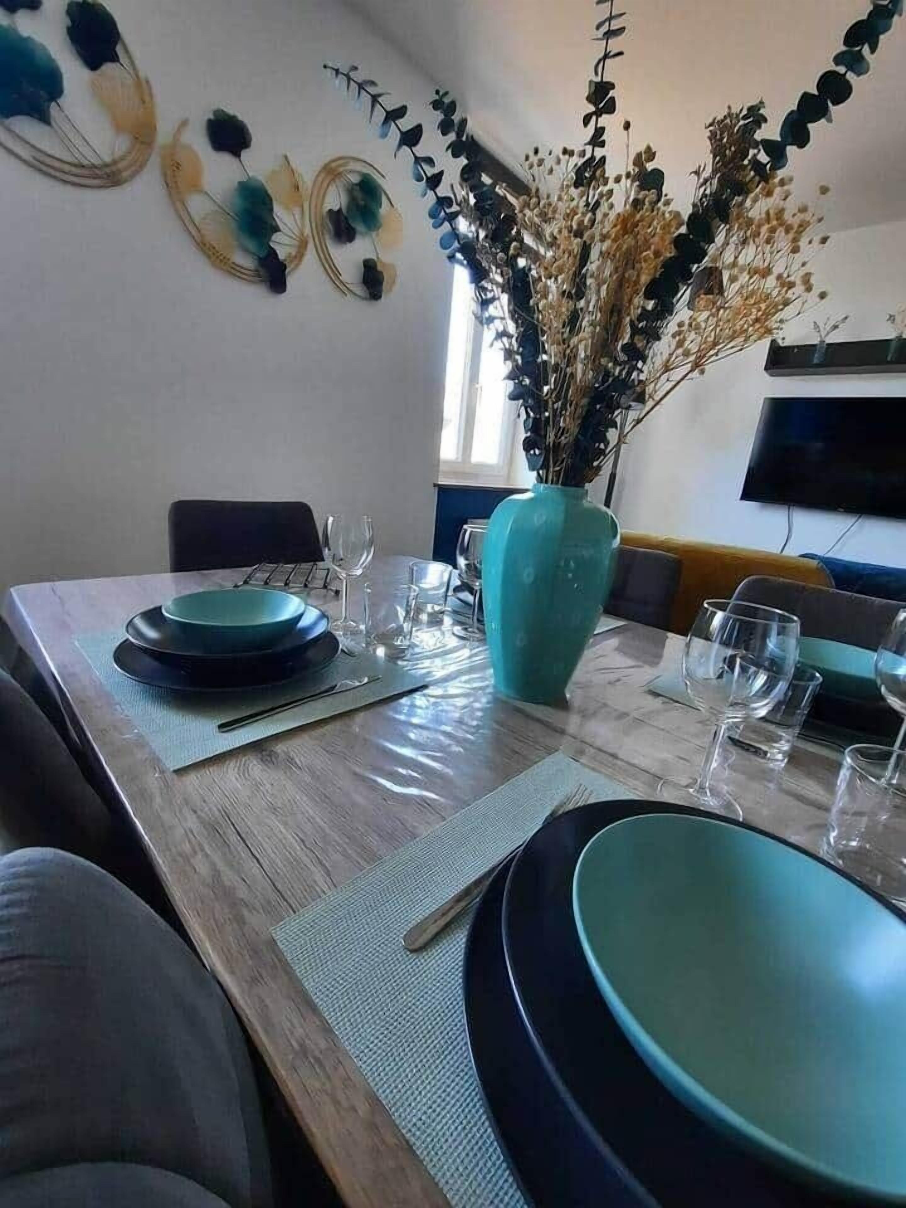 A dining area with a wooden table set for four, a vase with dried flowers, and a flat-screen TV mounted on the wall.