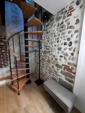 A spiral staircase with wooden steps and metal railings, a stone wall, and a bench. - Duplex Chic et Confort (Fécamp)