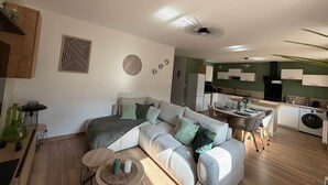 Uma sala de estar moderna com um sofá, mesa de centro e objetos decorativos. Uma cozinha com armários brancos e uma área de refeições com mesa e cadeiras são visíveis ao fundo.