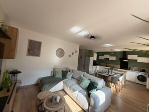 Ein modernes Wohnzimmer mit einer Couch, einem Couchtisch und Dekorationselementen. Im Hintergrund ist eine Küche mit weißen Schränken und ein Essbereich mit Tisch und Stühlen zu sehen.