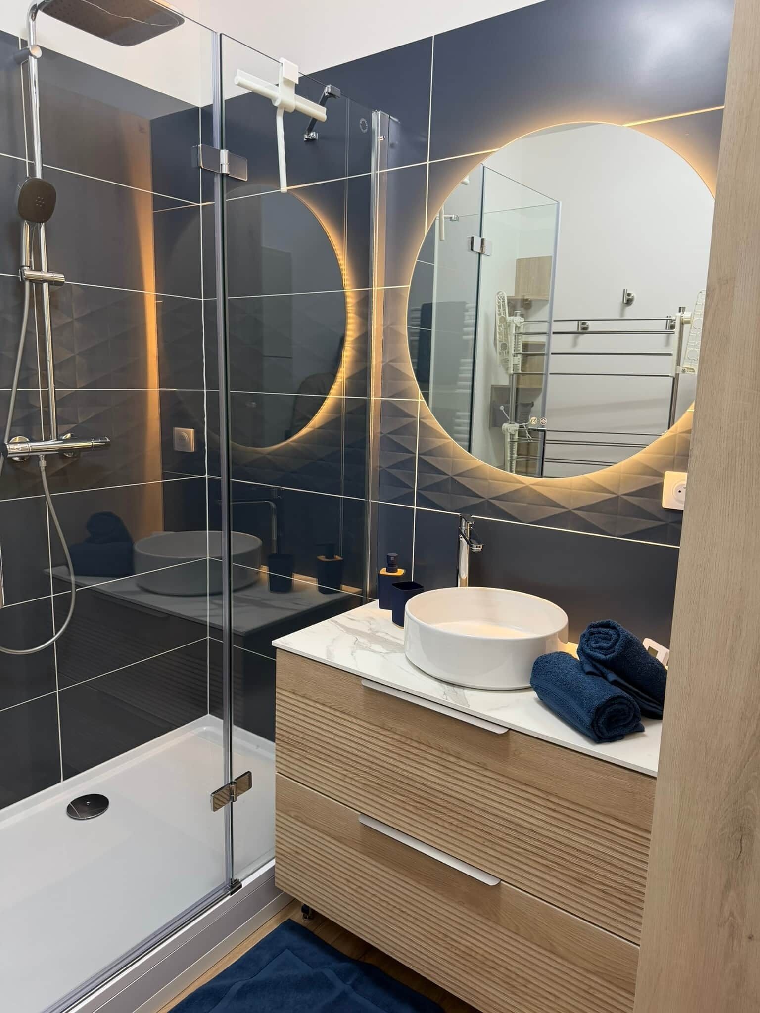 Ein modernes Badezimmer mit einer Duschkabine aus Glas, einem runden Spiegel, einem weißen Waschbecken und einer Holzeinrichtung.