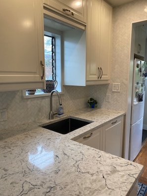 A kitchen with white cabinets, a marble countertop, a sink, and a window with a view. - Charming West Coast Retreat – Walk to Beach, Easy Access to FIFA World Cup (West Vancouver)