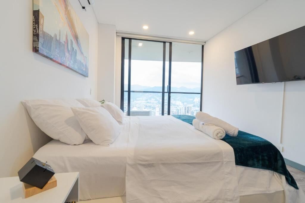 A hotel room with a bed, a flat-screen TV, a painting, and a window with a city view.