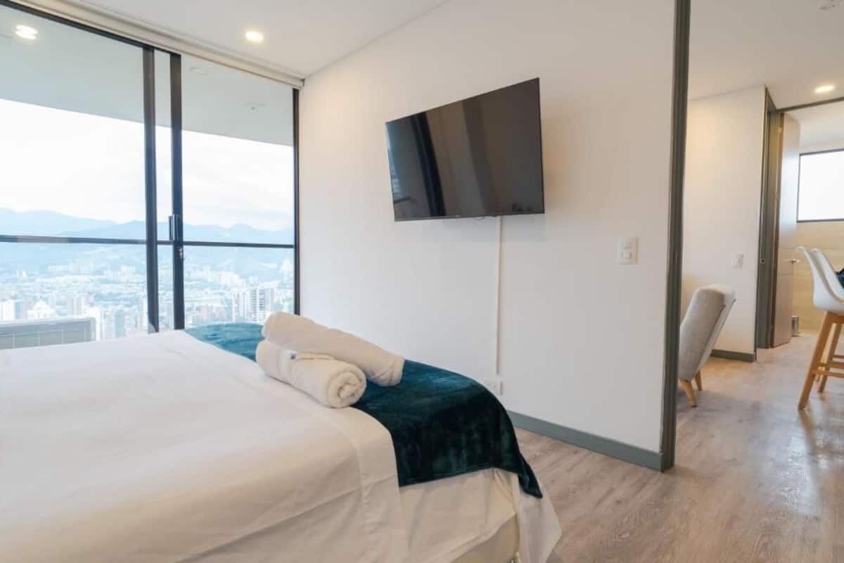 A modern hotel room with a bed, a flat-screen TV mounted on the wall, and a city view through large windows.