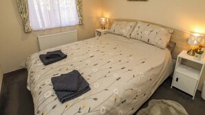 A bedroom with a bed, a nightstand, a lamp, a radiator, a window with curtains, and a painting on the wall. - Gardeners Lodge (Belford)