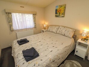 A bedroom with a bed, a nightstand, a lamp, a radiator, a window with curtains, and a painting on the wall. - Gardeners Lodge (Belford)