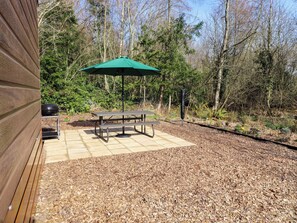 A patio with a picnic table and umbrella, surrounded by trees and a wooden deck. - Gardeners Lodge (Belford)