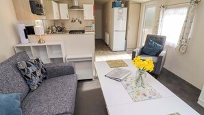 A modern living room with a grey sofa, a coffee table with a vase of yellow flowers, a grey armchair, and a view into the kitchen area. - Gardeners Lodge (Belford)