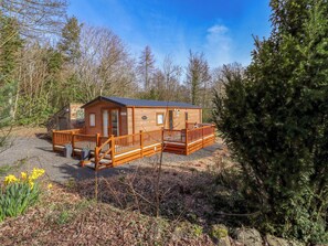 Une cabane en bois dotée d’une terrasse, entourée d’arbres et bordée d’un chemin de gravier.