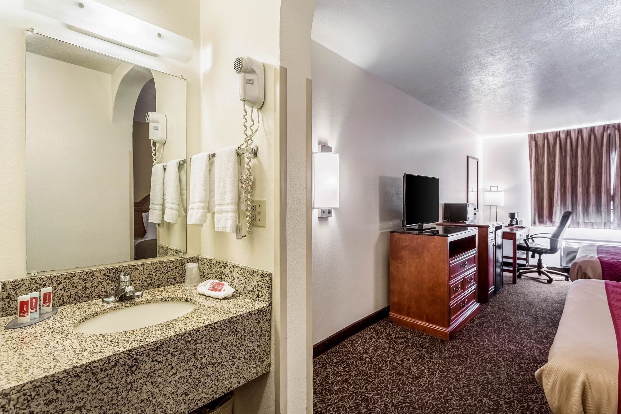 A hotel room with a bathroom, a desk, a TV, and a bed.