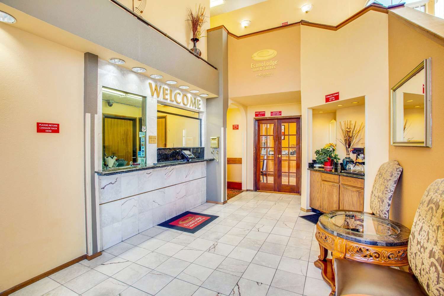 A hotel reception area with a marble counter, a 'Welcome' sign, a reception desk, a door, and a seating area with a glass table and chairs.