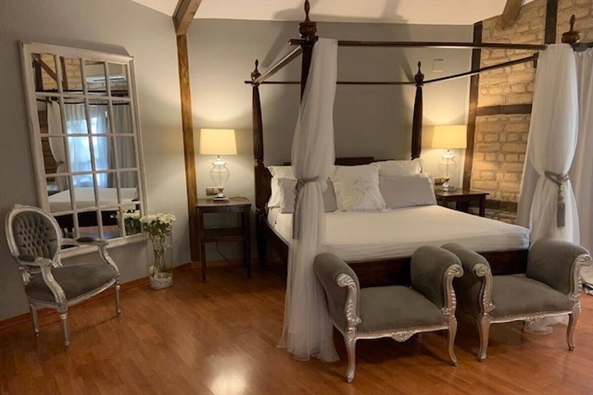 A four-poster bed with a canopy, two armchairs, a nightstand, a lamp, and a mirror with a window view.