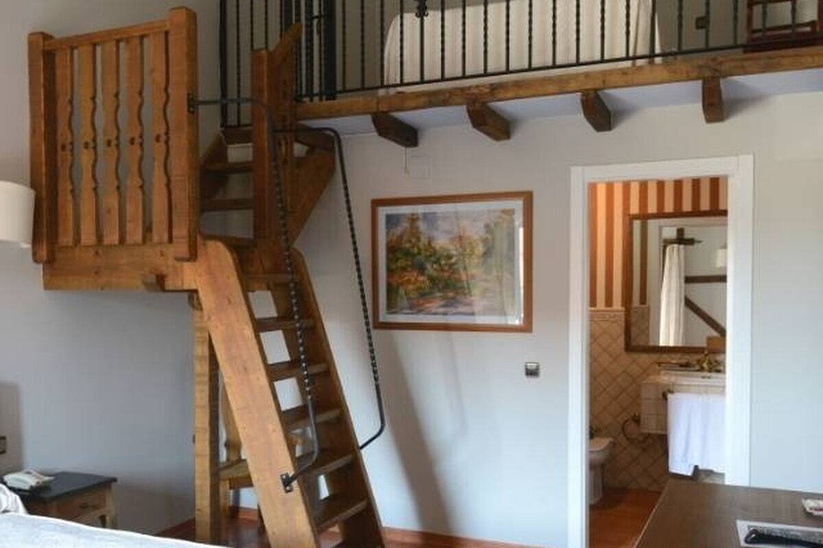 A room with a wooden staircase, a painting, a bathroom with a shower, and a bed.