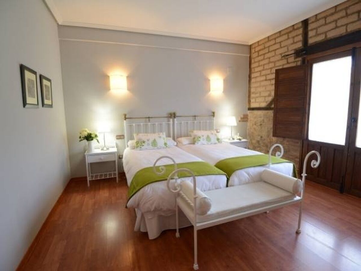 Una cama doble con estructura de metal, dos mesitas de noche con lámparas, una pared de ladrillo y un suelo de madera.
