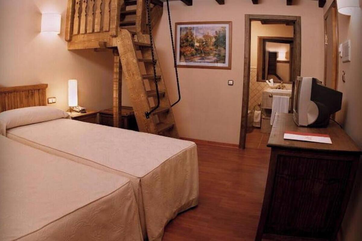 A bedroom with a bed, a desk with a computer, a painting, and a staircase.