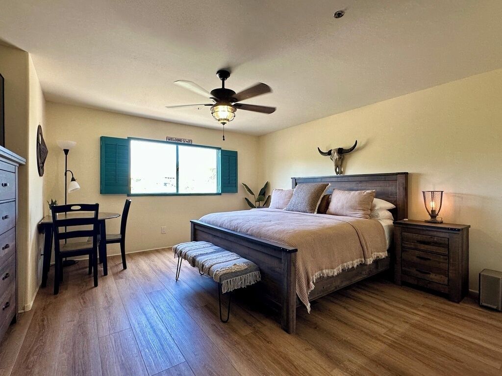 A bedroom with a bed, a desk, a chair, a bench, a bedside table, a lamp, a ceiling fan, and a wall-mounted bull skull decoration.