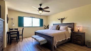 Quarto com cama, escrivaninha, cadeira, banco, mesa de cabeceira, abajur, ventilador de teto e uma caveira de boi presa à parede.