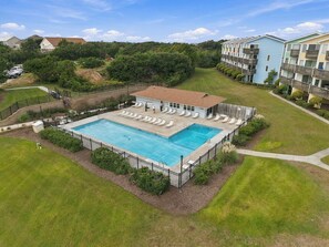 A swimming pool with lounge chairs, surrounded by a fence, and adjacent to a building with a red roof. - Welcome to Sound of the Sea 102N (Emerald Isle)