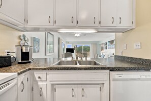 A kitchen with white cabinets, a granite countertop, a built-in dishwasher, and a view to the outdoors. - Welcome to Sound of the Sea 102N (Emerald Isle)