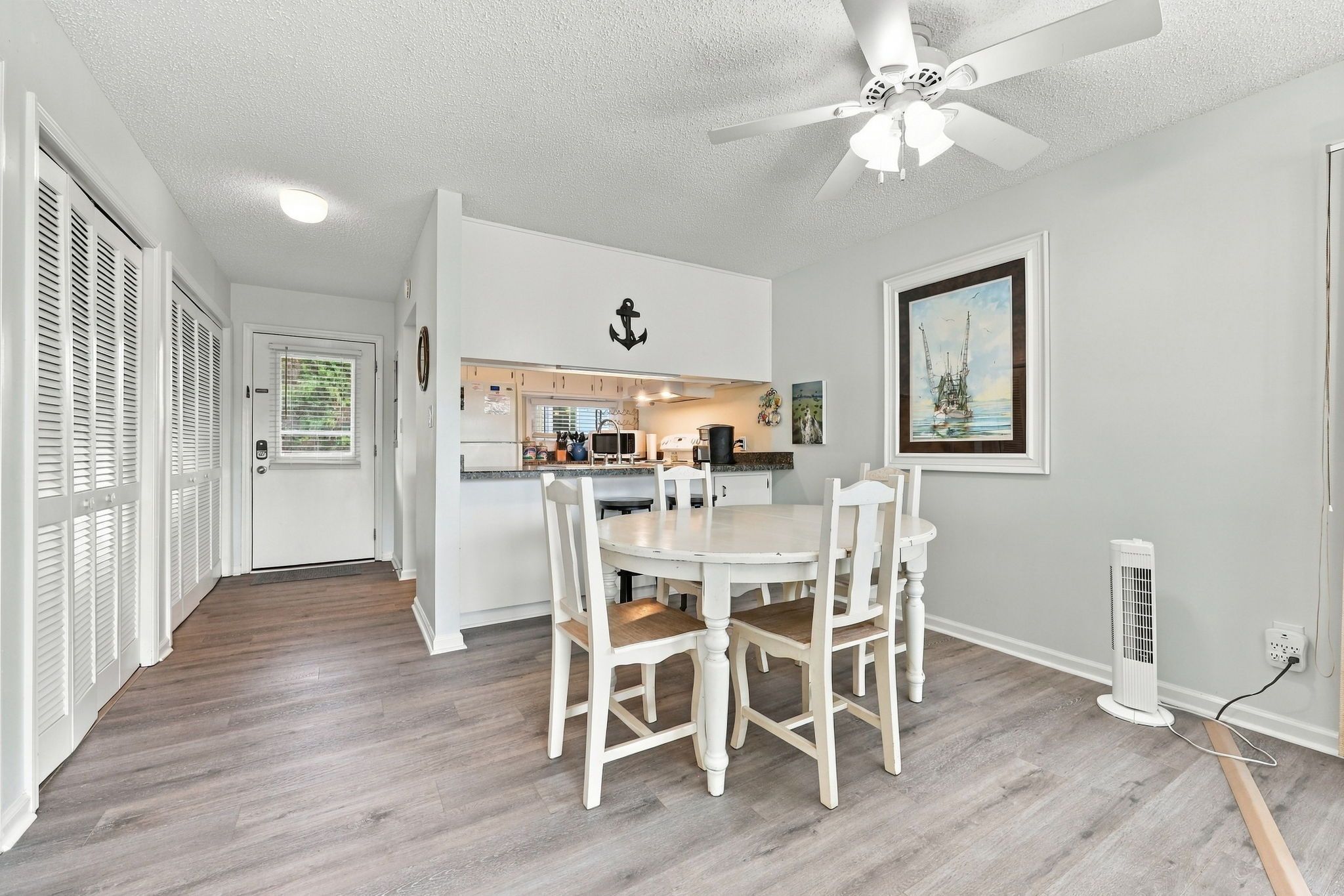 A kitchen with white cabinets, a dining table with chairs, a ceiling fan, and a framed painting on the wall.