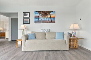 A living room with a sofa, two side tables, a wall-mounted lamp, and framed pictures. - Welcome to Sound of the Sea 102N (Emerald Isle)
