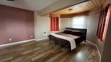 A bedroom with a wooden floor, a bed, a window with curtains, and a ceiling fan.