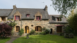 Un bâtiment en pierre doté de fenêtres en arc de cercle et d’une porte centrale, entouré d’une pelouse et d’un jardin bien entretenus.