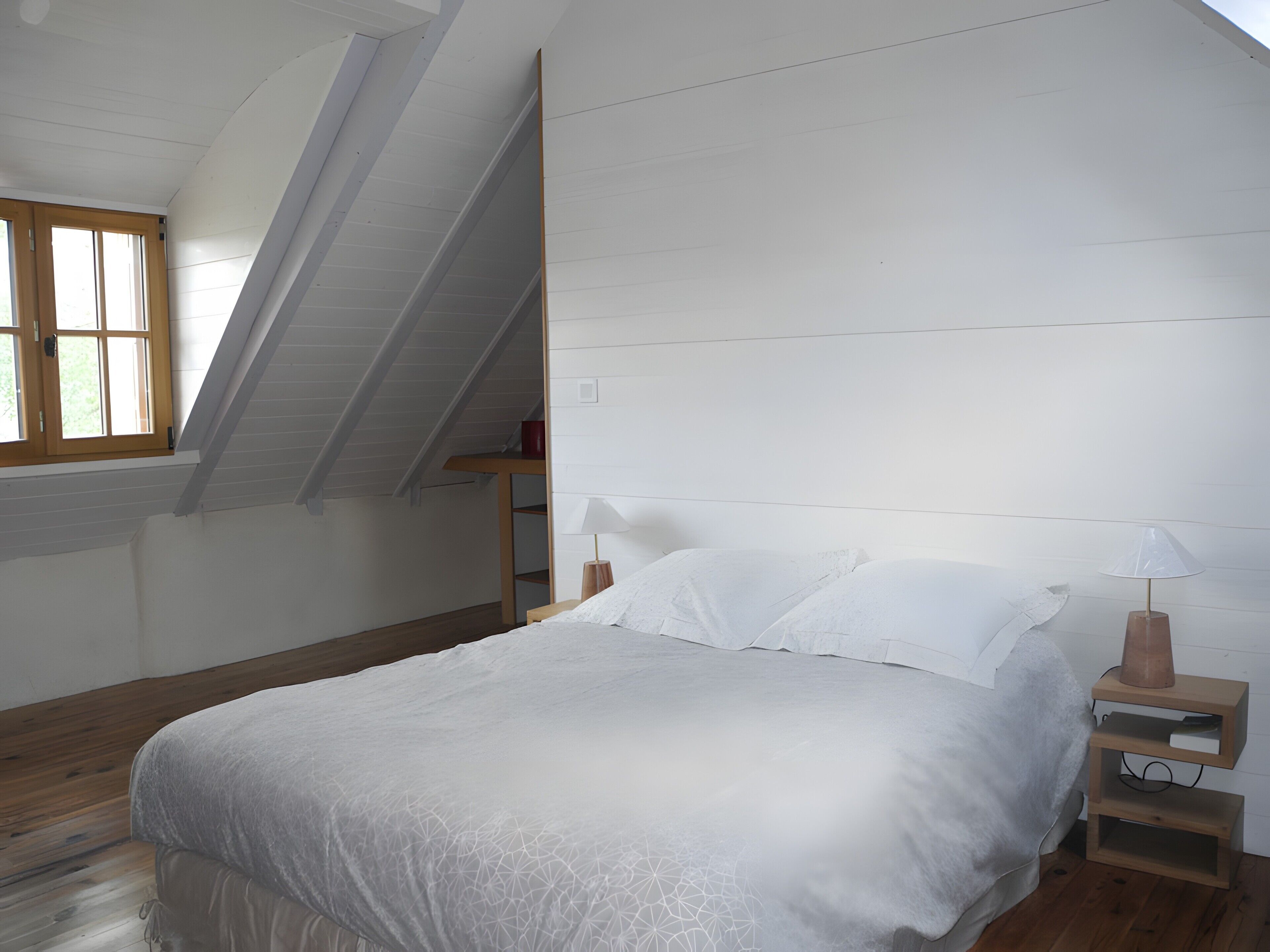 Une chambre avec un plafond en pente, un lit recouvert de draps blancs, deux tables de chevet avec des lampes et une fenêtre avec des cadres en bois.